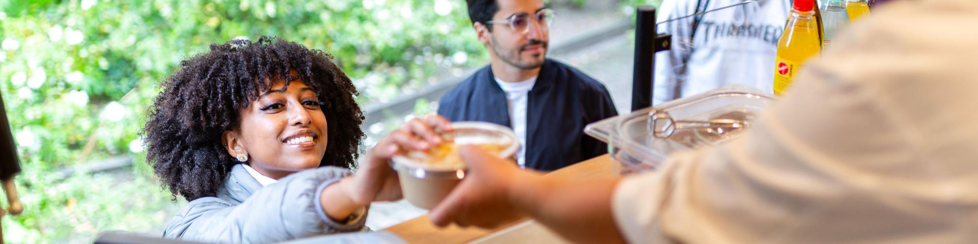 Foodtruckmitarbeiter*in gibt ein Essen in einer Schale mit Deckel über die Theke.