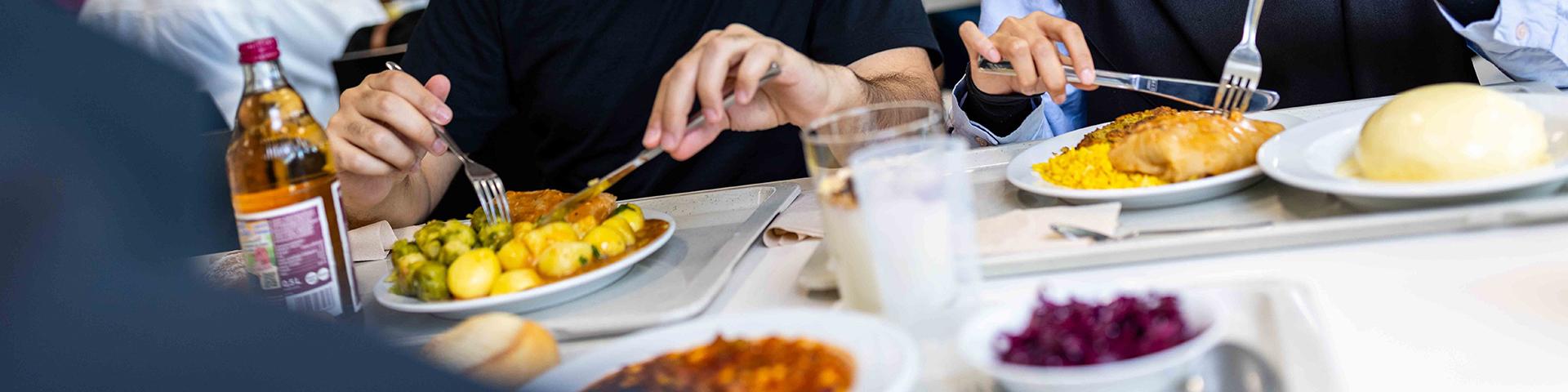 Studierende essen ihr Mensaessen, bestehend auf mehreren Komponenten und Getränken. 