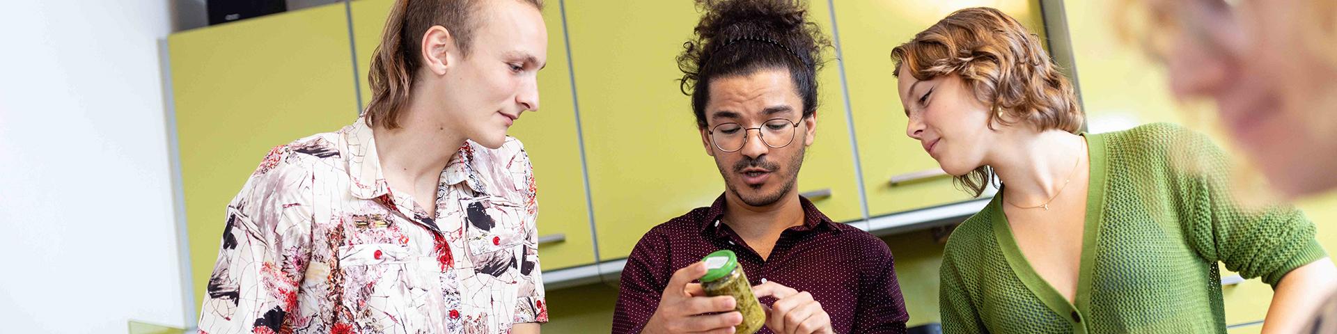 Studierende beim Kochen in einer Wohnheimküche. Zusammen schauen sie auf die Inhaltsstoffe ein Lebensmittels im Glas.