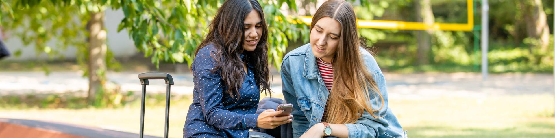 Zwei Studierende unterhalten sich miteinander und schauen dabei auf Smartphone. Eine Studierende hat einen Koffer dabei.
