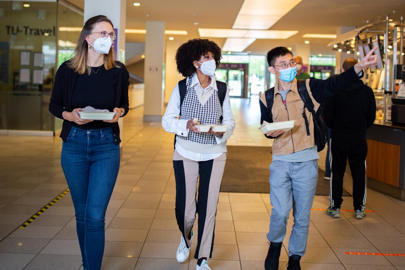 Studierende mit Maske in einem Mensa Foyer, eingeschweißtes Essen in der Hand haltend