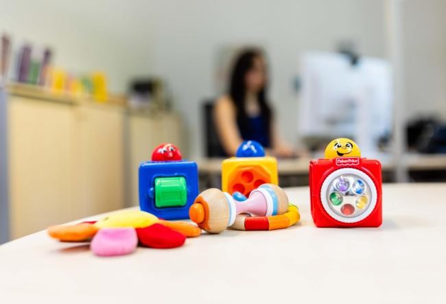 Kinderspielzeug auf einem Schreibtisch. Im Hintergrund sieht man eine Mutter am PC sitzend.