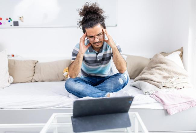 A stressed man sits on the bed and holds his head