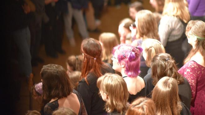 A group of singers can be seen. A person with purple hair in the middle of the crowd stands out.