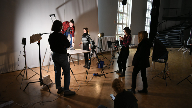Group of concentrated people standing in a circle around a simple filmset with a camera, a stativ and light sources, filming a sceen