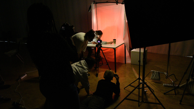 A dark room with a good lighted table in the middle. A camera is pointed on a little object on top of the table and four people observe the scene critically.