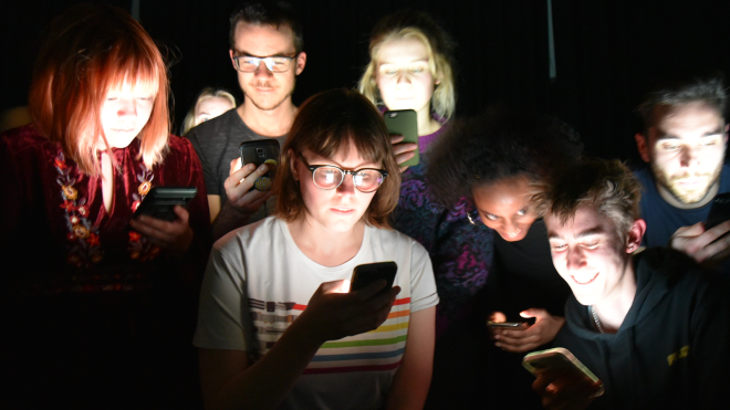 A group of diverse people stand close together in a dark environment, all looking at their own brightly lit cell phones. The faces, all showing different emotions, are illuminated only by the smartphones.