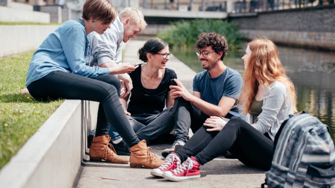 The picture shows the young people sitting next to a river and talking with a smile.