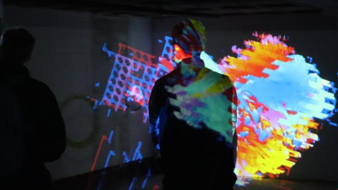 Person with short hair stands in an art exhibition, only the back and the back of the head are visible. The person is immersed in the colours. The picture seems intense and a bit thoughtful.
