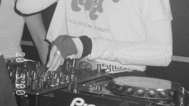 A DJ mixing at a console, wearing a printed T-shirt and fingerless gloves. Black-and-white club photo.