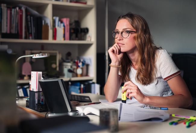 Young woman wearing glasses is studying at a desk with books, a laptop, and highlighters