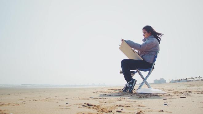 A person is sitting on a chair at the beach, holding a large clipboard.