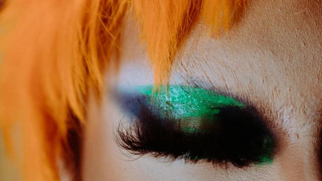A close-up of a person with intensely orange hair, shimmering green eyeshadow, and a dramatic expression. Their eyes are closed.