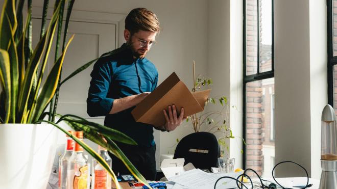 A person is standing in front of a messy desk holding a folder in their hands.